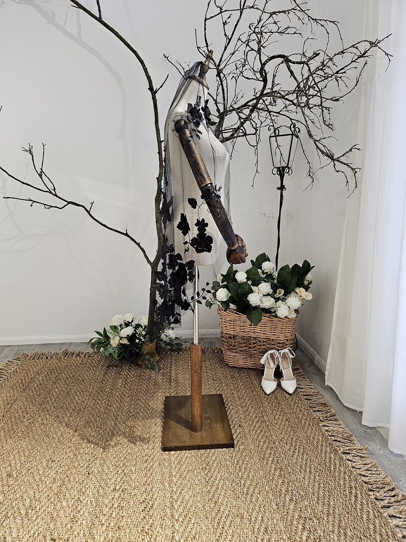 K&ouml;nnte beinhalten: Eine Schaufensterpuppe mit einem transparenten Schleier mit schwarzen Blumenstickereien. Die Schaufensterpuppe steht auf einem Holzsockel, mit einem Korb mit wei&szlig;en Blumen und einem Paar wei&szlig;er Abs&auml;tze. Die Szene spielt sich auf einem gewebten Teppich mit Fransen ab.