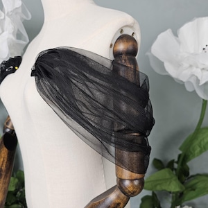 Puede incluir: Detalle de manga de tul negro en un maniquí. La manga está drapeada y fruncida, creando un efecto de capas. El maniquí es de color crema, con brazos de madera y una base de madera. El fondo incluye acentos florales blancos.