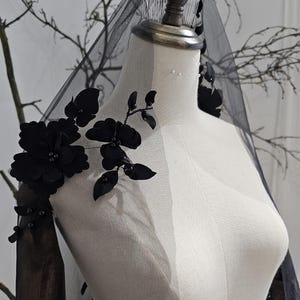 K&ouml;nnte beinhalten: Ein schwarzer Brautschleier mit floralen Verzierungen. Der Schleier besteht aus durchsichtigem Stoff und weist gro&szlig;e, dunkle Bl&uuml;ten und Bl&auml;tter auf. Der Schleier wird auf einer Schaufensterpuppe pr&auml;sentiert.