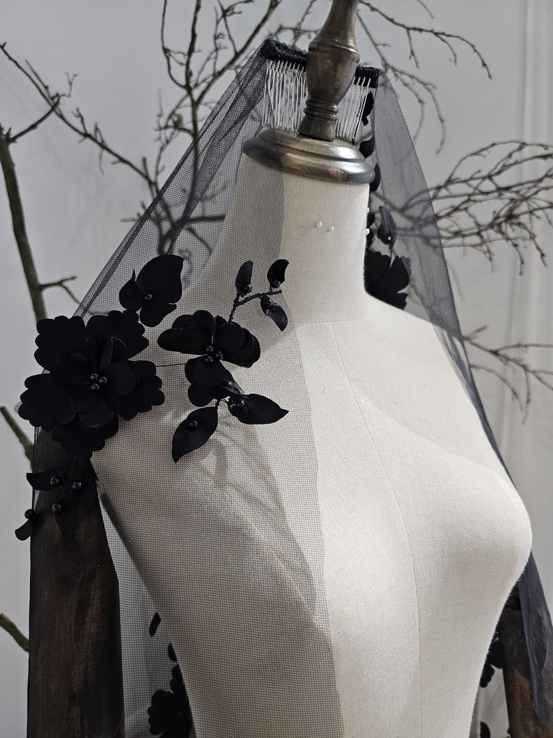 K&ouml;nnte beinhalten: Ein schwarzer Brautschleier mit floralen Verzierungen. Der Schleier besteht aus durchsichtigem Stoff und weist gro&szlig;e, dunkle Bl&uuml;ten und Bl&auml;tter auf. Der Schleier wird auf einer Schaufensterpuppe pr&auml;sentiert.