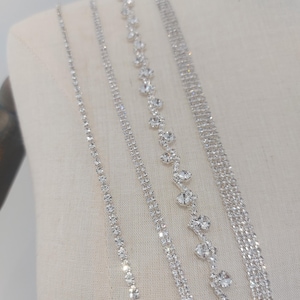 Puede incluir: Cinco tiras de ribete de strass plateado con diferentes tamaños y patrones de strass. El ribete se muestra sobre un fondo de tela blanca.