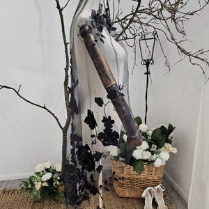 K&ouml;nnte beinhalten: Ein schwarzer T&uuml;llschleier mit 3D-Blumenschmuck, drapiert auf einer Schaufensterpuppe. Die floralen Elemente sind an den R&auml;ndern konzentriert und flie&szlig;en den Schleier hinunter. Die Schaufensterpuppe steht auf einem Holzst&auml;nder, mit einem Weidenkorb mit wei&szlig;en Blumen und wei&szlig;en Abs&auml;tzen im Hintergrund.