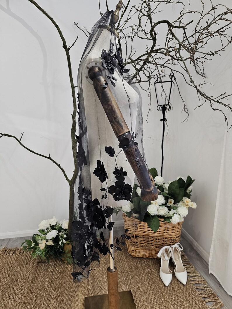 K&ouml;nnte beinhalten: Ein schwarzer T&uuml;llschleier mit 3D-Blumenschmuck, drapiert auf einer Schaufensterpuppe. Die floralen Elemente sind an den R&auml;ndern konzentriert und flie&szlig;en den Schleier hinunter. Die Schaufensterpuppe steht auf einem Holzst&auml;nder, mit einem Weidenkorb mit wei&szlig;en Blumen und wei&szlig;en Abs&auml;tzen im Hintergrund.