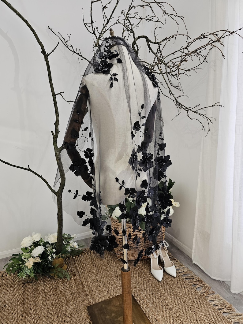 K&ouml;nnte beinhalten: Ein schwarzer T&uuml;llschleier mit floralen Verzierungen ist auf einer Schaufensterpuppe drapiert. Der Schleier ist mit dunklen floralen Akzenten versehen, die einen dramatischen Effekt erzeugen. Die Schaufensterpuppe steht in der N&auml;he eines Korbs mit wei&szlig;en Blumen und wei&szlig;en Abs&auml;tzen.