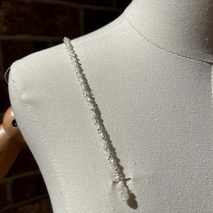 May include: A delicate silver-toned chain necklace featuring small, evenly spaced white beads along its length. The necklace rests against a neutral-colored mannequin torso, showcasing a simple, linear design with a small, dangling bead element at the end.