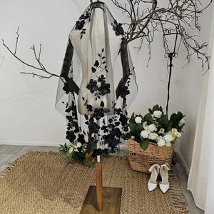K&ouml;nnte beinhalten: Ein schwarzer T&uuml;llschleier mit floralen Verzierungen wird auf einer Schaufensterpuppe pr&auml;sentiert. Der Schleier hat aufwendige schwarze Blumenapplikationen und ist &uuml;ber die Schultern der Schaufensterpuppe drapiert. Die Szene umfasst einen Weidenkorb mit wei&szlig;en Blumen und ein Paar wei&szlig;e Abs&auml;tze.
