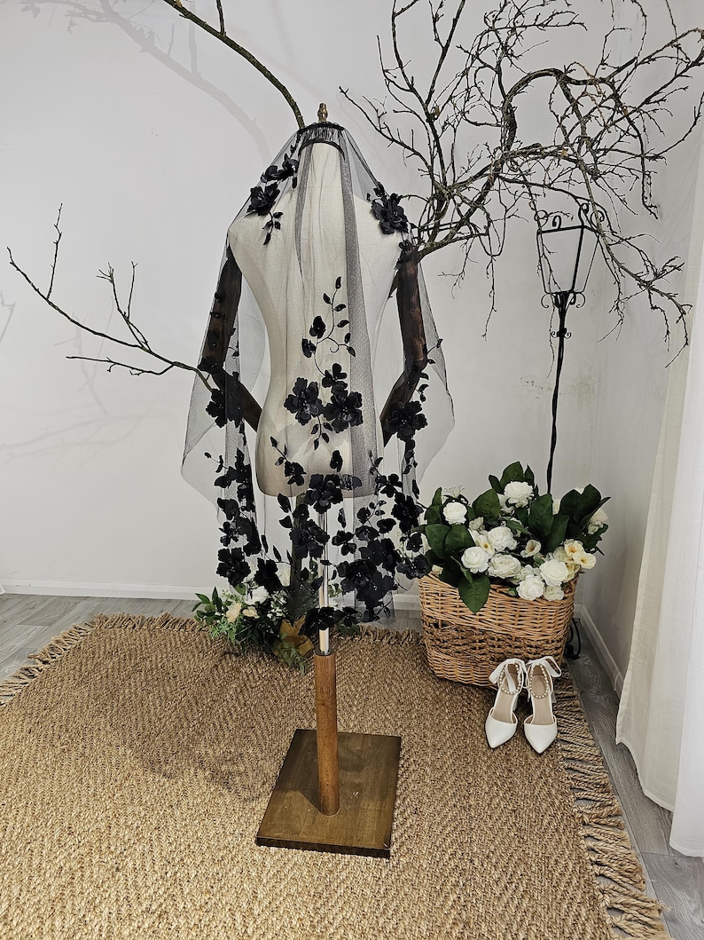 K&ouml;nnte beinhalten: Ein schwarzer T&uuml;llschleier mit floralen Verzierungen wird auf einer Schaufensterpuppe pr&auml;sentiert. Der Schleier hat aufwendige schwarze Blumenapplikationen und ist &uuml;ber die Schultern der Schaufensterpuppe drapiert. Die Szene umfasst einen Weidenkorb mit wei&szlig;en Blumen und ein Paar wei&szlig;e Abs&auml;tze.