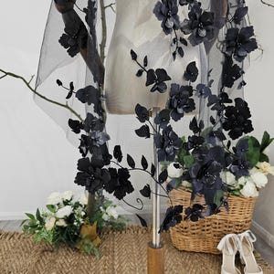 K&ouml;nnte beinhalten: Schwarzer floraler Spitzen-Schleier mit 3D-Blumenverzierungen. Der Schleier ist &uuml;ber einer Schaufensterpuppe drapiert und zeigt das aufwendige Design. Die Blumen sind entlang der R&auml;nder und &uuml;ber dem transparenten Stoff angeordnet und schaffen einen romantischen und eleganten Look. F&uuml;r Brautmode.