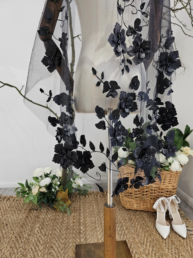 K&ouml;nnte beinhalten: Schwarzer floraler Spitzen-Schleier mit 3D-Blumenverzierungen. Der Schleier ist &uuml;ber einer Schaufensterpuppe drapiert und zeigt das aufwendige Design. Die Blumen sind entlang der R&auml;nder und &uuml;ber dem transparenten Stoff angeordnet und schaffen einen romantischen und eleganten Look. F&uuml;r Brautmode.
