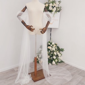 May include: A white tulle bridal cape with long, flowing fabric attached to a dress form. The dress form has wooden arms and is set on a wooden stand. A bouquet of white flowers is in the background.