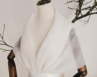 Puede incluir: Un chal blanco y transparente drapeado sobre un maniquí. El chal tiene un escote en V profundo y se asegura con una banda blanca en la cintura. El maniquí tiene brazos de madera y un cuello de madera. El fondo es de color neutro.