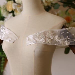 May include: A white off-the-shoulder lace bridal sash with delicate floral embroidery.