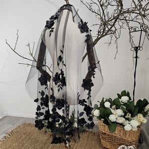 K&ouml;nnte beinhalten: Ein schwarzer T&uuml;llschleier mit floralen Verzierungen. Der Schleier wird auf einer Schaufensterpuppe pr&auml;sentiert, die die filigranen Blumendetails zeigt. Die Blumen sind an den R&auml;ndern angeordnet und fallen den Schleier hinunter. Im Hintergrund ein Korb mit wei&szlig;en Blumen und wei&szlig;e Schuhe.