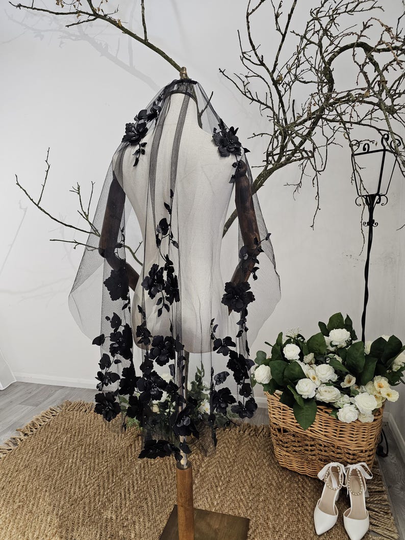 K&ouml;nnte beinhalten: Ein schwarzer T&uuml;llschleier mit floralen Verzierungen. Der Schleier wird auf einer Schaufensterpuppe pr&auml;sentiert, die die filigranen Blumendetails zeigt. Die Blumen sind an den R&auml;ndern angeordnet und fallen den Schleier hinunter. Im Hintergrund ein Korb mit wei&szlig;en Blumen und wei&szlig;e Schuhe.