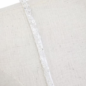 May include: A thin, white spaghetti strap adorned with clear, faceted glass beads that catch the light. The strap is positioned diagonally across a textured, off-white fabric surface, showcasing the intricate, sparkling beadwork detail.