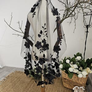 K&ouml;nnte beinhalten: Ein schwarzer Brautschleier mit floralen Verzierungen. Der Schleier wird auf einer Schaufensterpuppe pr&auml;sentiert, die die detaillierten Blumendesigns hervorhebt. Die Blumen sind gruppiert und &uuml;ber dem transparenten Stoff verteilt, was einen romantischen und eleganten Look erzeugt. Ein Hochzeitsaccessoire.