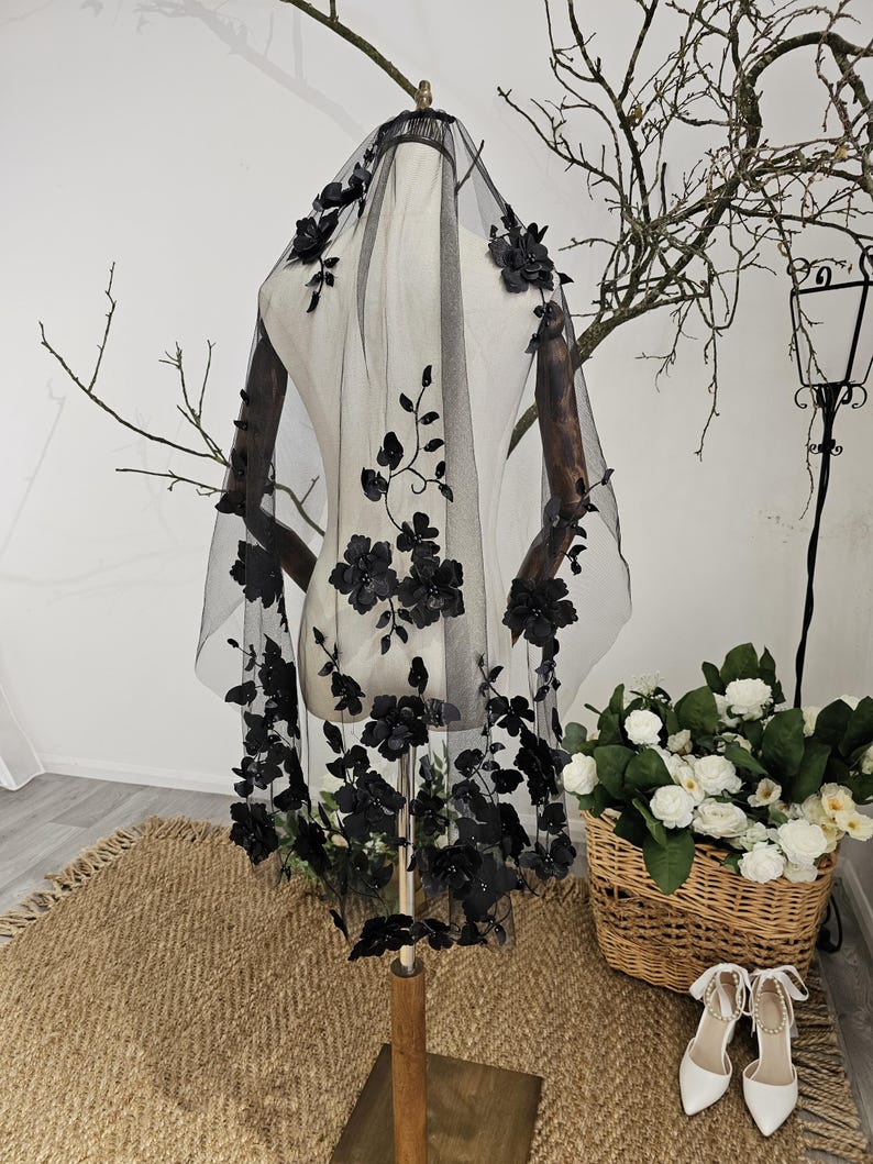 K&ouml;nnte beinhalten: Ein schwarzer Brautschleier mit floralen Verzierungen. Der Schleier wird auf einer Schaufensterpuppe pr&auml;sentiert, die die detaillierten Blumendesigns hervorhebt. Die Blumen sind gruppiert und &uuml;ber dem transparenten Stoff verteilt, was einen romantischen und eleganten Look erzeugt. Ein Hochzeitsaccessoire.