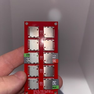 May include: A red SD card reader with eight slots, each holding a silver SD card. The reader has green and white labels with the number 32. Text on the reader includes "Made By Justin W. Burris" and "Not Final Product."