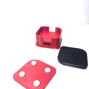 May include: A red square coaster holder with a black coaster and a red coaster with four white circular pads. The holder is square with a slot for the coasters. The coasters are made of a smooth material.