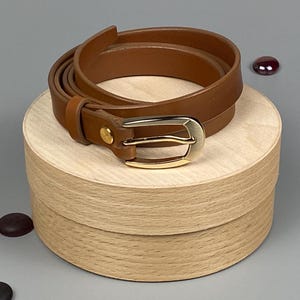 May include: A brown leather belt with a gold-colored buckle is coiled on top of a round wooden box. The belt is approximately 1 inch wide. The background is a solid gray color.