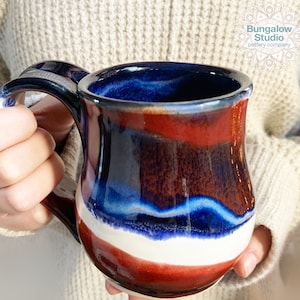 May include: A handmade ceramic mug with a patriotic color scheme. The mug features bands of red, white, and blue glaze, with a dark blue interior and handle. The mug is held by a person wearing a cream-colored sweater. The mug has the text "Bungalow Studio pottery company".
