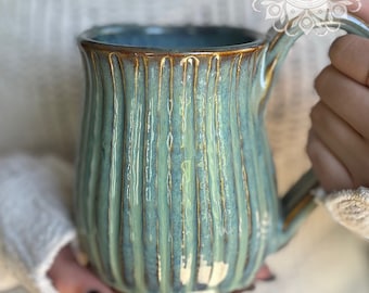 May include: A handmade ceramic mug with a ribbed design in shades of teal and blue. The mug has a brown rim and handle, and the text "Bungalow Studio pottery company" is visible. The mug is held by a person.