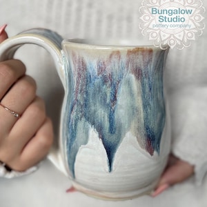 May include: A handmade ceramic mug with a unique glaze. The mug features a white base with blue, red, and cream-colored drips. The mug has a large handle and the text "Bungalow Studio pottery company" is visible.