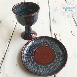 May include: Handmade ceramic chalice and plate set. The chalice has a wide rim and a pedestal base. The plate is round. Both pieces feature a glossy glaze with shades of brown, blue, and green. The set is from Bungalow Studio pottery company.