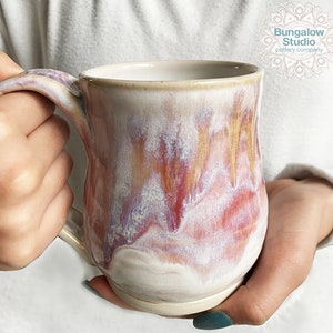 May include: A handmade ceramic mug with a unique glaze. The mug features a cream-colored base with flowing patterns of red, orange, and purple. The handle is integrated into the design. The mug is from Bungalow Studio pottery company.
