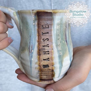 May include: Handmade ceramic mug with a unique glaze in shades of white, blue, and brown. The mug features the word "NAMASTE" vertically on a brown band. The mug has a large handle and is held by a person.