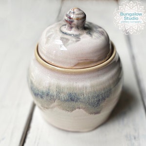May include: A ceramic jar with a lid, featuring a light pink and blue glaze. The jar has a rounded shape with a lid and a small knob on top. The jar is handmade and has a unique, artistic appearance.
