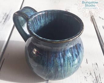 Può includere: Una tazza in ceramica fatta a mano con una glassa blu scuro e verde. La tazza ha un corpo arrotondato, un manico curvo e un bordo dorato. La tazza è di Bungalow Studio pottery company.