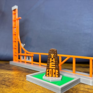 May include: A miniature orange and gray roller coaster model with a brown sign that reads "THE EDGE". The model has a tall vertical lift hill and a curved track section. The base is gray with a green patch.