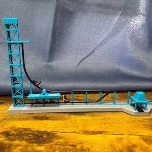 May include: A miniature roller coaster model with a teal tower, track, and supports. The track is black, and the base is gray. The model is set against a blue backdrop and a wooden surface.