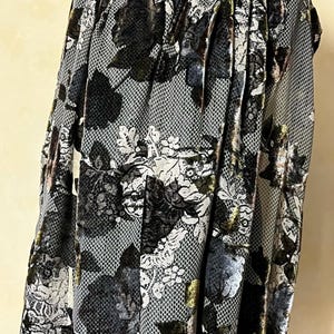 May include: A draped textile featuring a floral pattern in black, white, and olive green. The fabric has a net-like overlay, creating a textured appearance. The design is a mix of dark and light tones, with a velvet-like sheen.