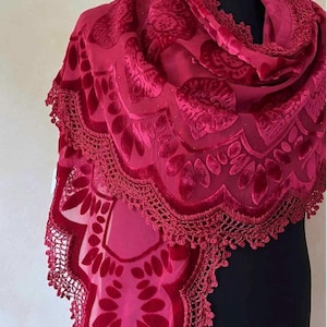 May include: A deep red velvet shawl featuring a semi-sheer base with a raised floral and leaf pattern. The edges are finished with a detailed scalloped crochet lace trim that follows the contour of the fabric, creating a textured border.