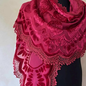 May include: A burgundy scarf with a floral velvet pattern and a crocheted lace trim. The scarf is draped over a black mannequin, showcasing its intricate design and rich color. The fabric appears soft and luxurious.