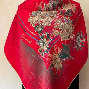 May include: A vibrant red scarf with a floral design in shades of beige, green, and blue. The scarf features the signature "Salimbeni" in cursive script. The edges are trimmed with a gold border.