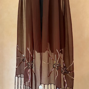 Pode incluir: Um lenço retangular castanho-escuro, feito de tecido leve e semitransparente. As bordas inferiores apresentam franjas longas e bordados florais decorativos com lantejoulas iridescentes. Uma pequena etiqueta branca está fixada na bainha.