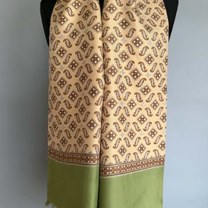 May include: A long scarf featuring a beige background with a repeating brown pattern of small squares and paisley-like shapes. The scarf has a wide solid olive green border at the bottom edge, finished with a delicate fringe along the hemline.