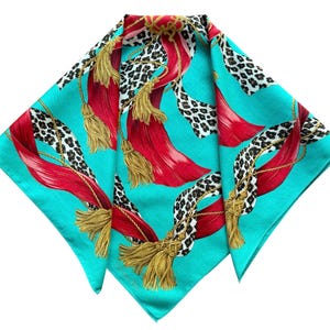 May include: A square scarf featuring a vibrant turquoise background with a repeating pattern of red ribbons, gold tassels, and black and white leopard print sections. The design includes thin gold rope accents intertwined with the ribbons and tassels.