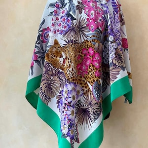 Op de afbeelding: Een vierkante zijden sjaal met een opvallend ontwerp van een luipaard omringd door kleurrijke bloemen en bladeren. De sjaal heeft een witte achtergrond met roze, paarse en groene accenten en een groene rand. De sjaal is gemaakt van licht materiaal.