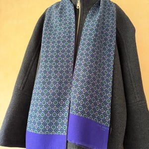 May include: A Ted Baker scarf featuring a repeating geometric pattern in shades of blue, green, and purple, finished with a solid purple border and fringe at the ends. The scarf is draped over a dark gray zip-up jacket with a high collar.