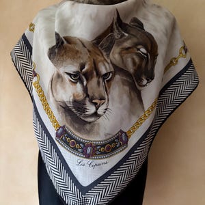Puede incluir: Un pañuelo cuadrado de seda con dos pumas marrones centrados sobre un fondo crema. El diseño incluye un motivo de cadena dorada, un collar decorativo de joyas moradas y doradas, y un borde de espiga en blanco y negro. El texto Les Copains está impreso.