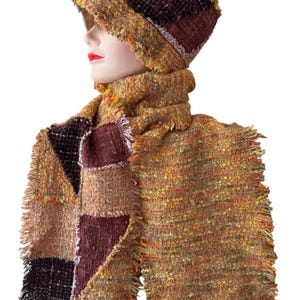 Puede incluir: Un conjunto de sombrero y bufanda texturizados con un diseño de patchwork en tonos amarillo mostaza, naranja quemado y marrón oscuro. El sombrero tiene un ala corta y la bufanda muestra un acabado de bordes deshilachados con tejido multicolor.
