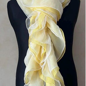 Könnte beinhalten: Ein langer, leichter Schal mit Lagen aus gerüschtem Stoff in abwechselnden Farbtönen von Blassgelb und Weiß. Der Schal hat eine transparente, geknitterte Textur und ist um eine schwarze Schaufensterpuppe drapiert, mit einem Etikett unten.