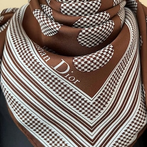 May include: A brown silk scarf with a white houndstooth and striped pattern. The scarf features the text "Christian Dior" in white. The scarf is folded, showcasing the intricate design and luxurious fabric.