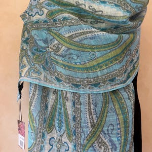 May include: A paisley-patterned scarf in shades of blue, green, and gray. The scarf is draped over a black form, showcasing the intricate design. The fabric appears to be lightweight, suitable for warmer weather.
