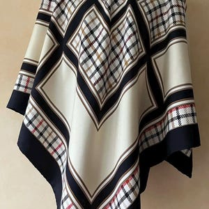 May include: A square scarf with a geometric pattern. The scarf features a cream-colored background with black, brown, and red plaid squares. The edges are trimmed with black and brown stripes.