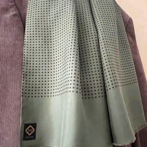 Op de afbeelding: Lichtgroene sjaal met een herhalend patroon van kleine donkere vierkantjes. De sjaal is gedrapeerd over een paarse corduroy blazer. De sjaal heeft een gefranjerde rand en een label met het woord "Pain".